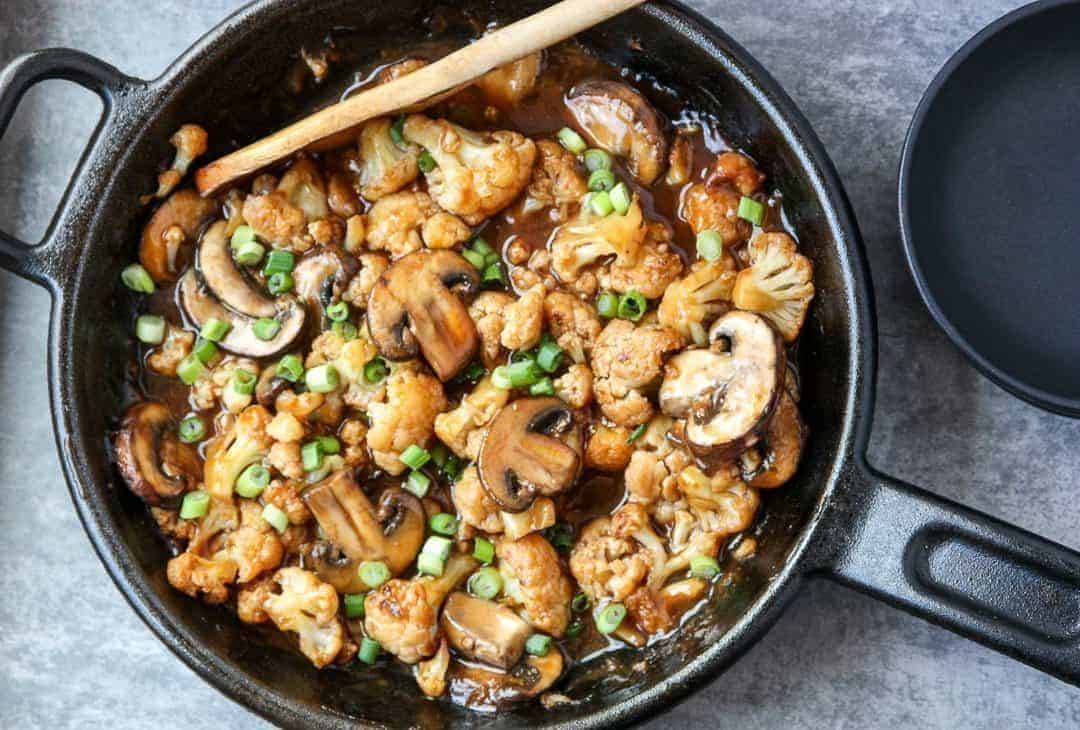 A skillet with mushrooms and cauliflower is stirred with a wooden spoon