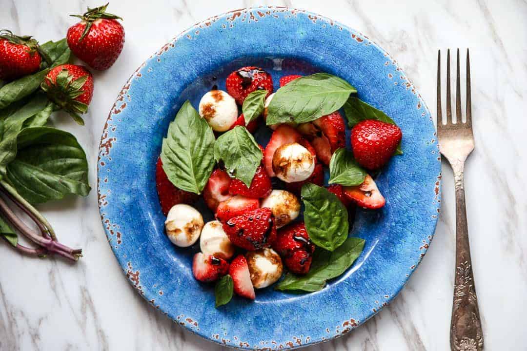 Top shot of basil, strawberries and mozzarella cheese balls on blue plate.