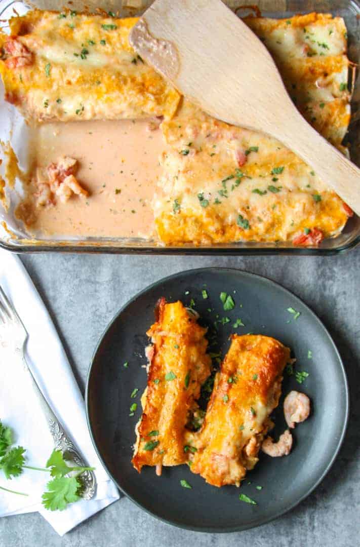 Two seafood enchiladas on a black plate beside a pan of seafood enchiladas