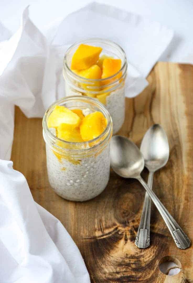 2 jars of tropical chia seed pudding topped with mango & pineapple chunks, on a board with a white napkins and two spoons