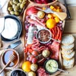 An irregularly shaped wooden charcuterie board filled with meat, cheese, olives, nuts and fruit, surrounded by crackers and bread.