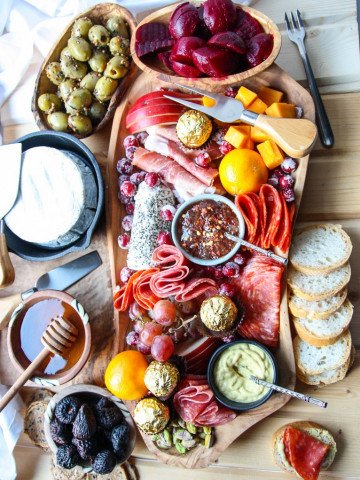 An irregularly shaped wooden charcuterie board filled with meat, cheese, olives, nuts and fruit, surrounded by crackers and bread.
