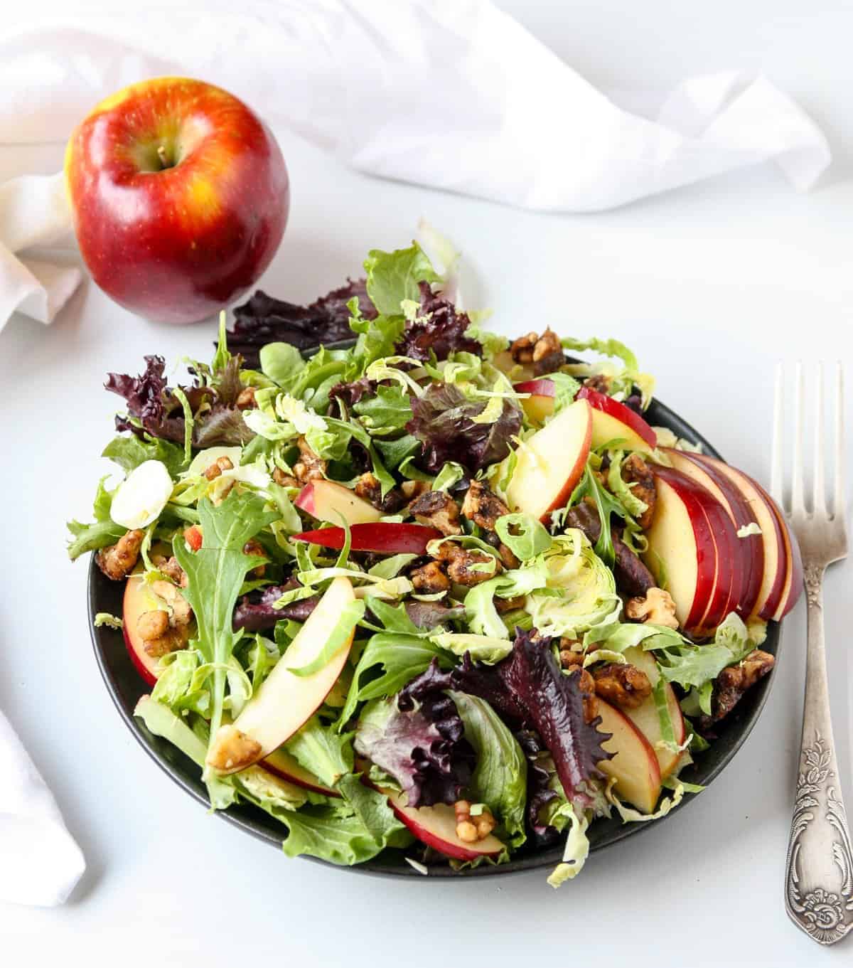 An apple and a plateful of salad on a white table with a fork.