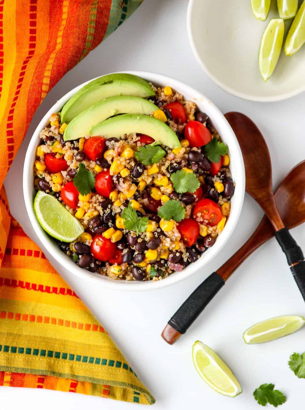A bowl of salad on a white table with two wooden salad spoons and a small dish of lime wedges.