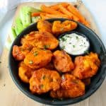 Gluten-free baked buffalo cauliflower bites on a black plate with carrot and celery sticks.