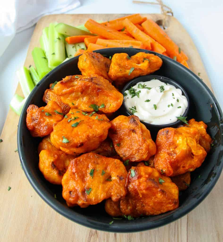 A black plate full of buffalo cauliflower bites with a small dish of ranch dip and carrot and celery sticks.