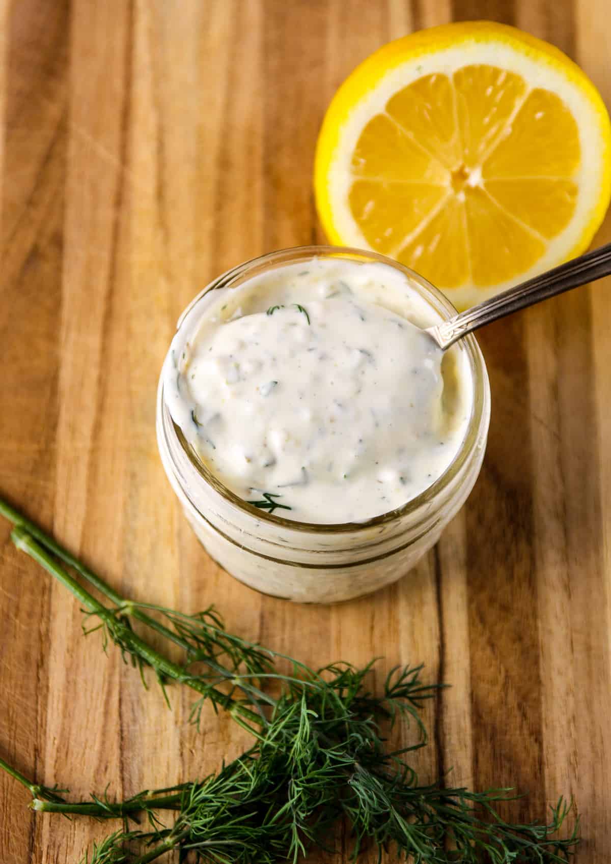 A jar of homemade tartar sauce with a lemon and a sprig of dill on a wooden board.
