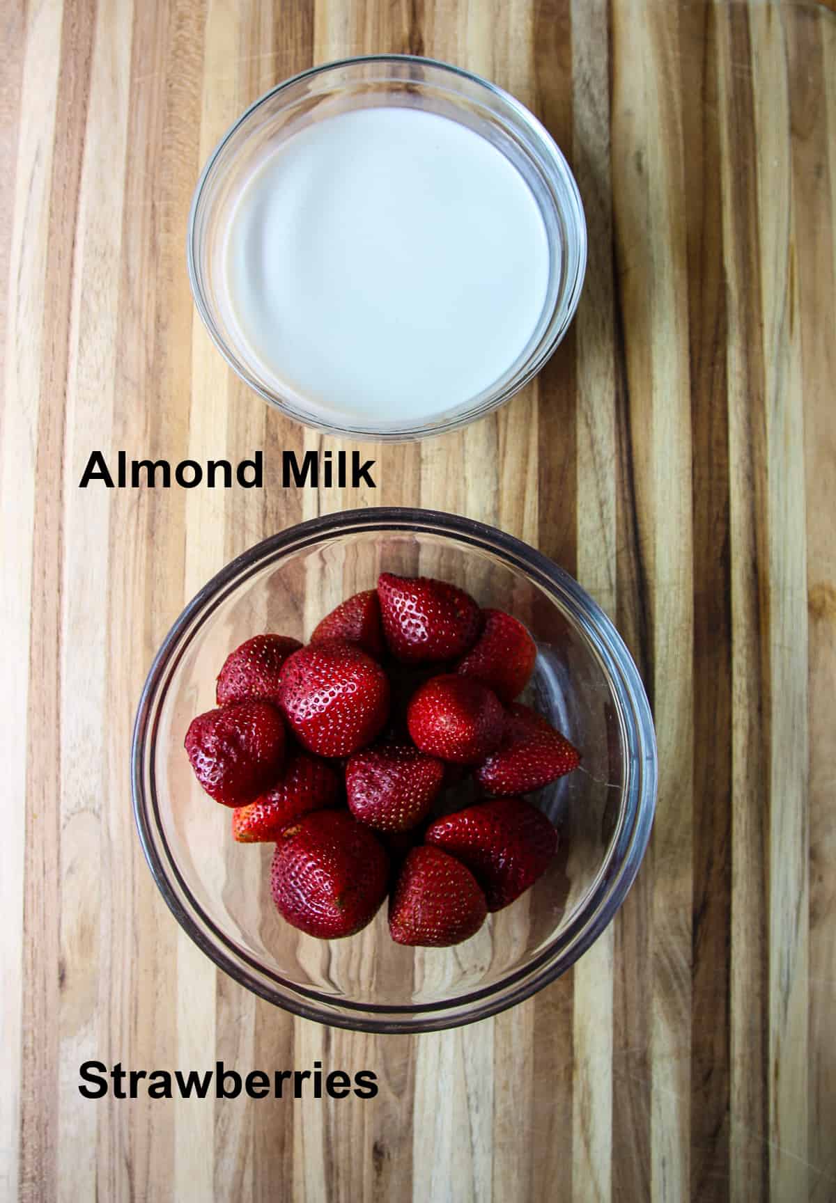 The two ingredients used to make strawberry popsicles; almond milk and strawberries, in glass bowls.