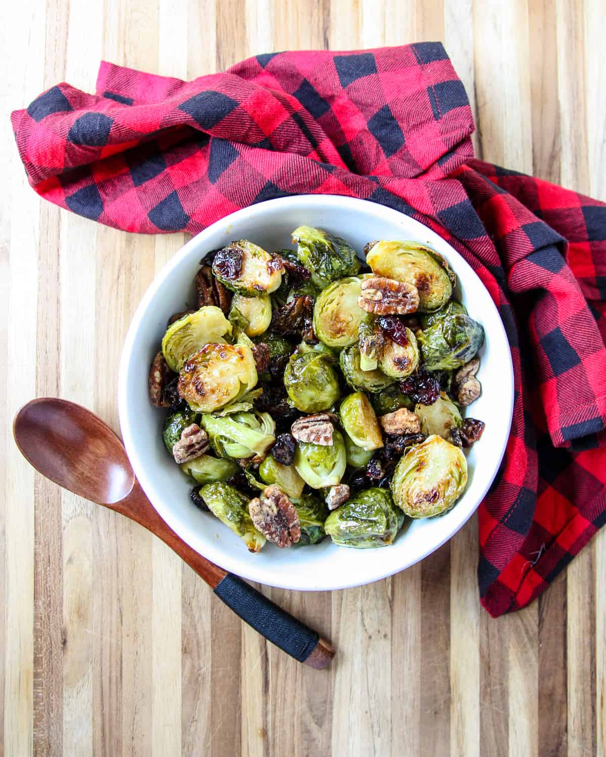 Brown sugar Brussels sprouts in a white bowl, a wooden spoon, and a black & red napkin.