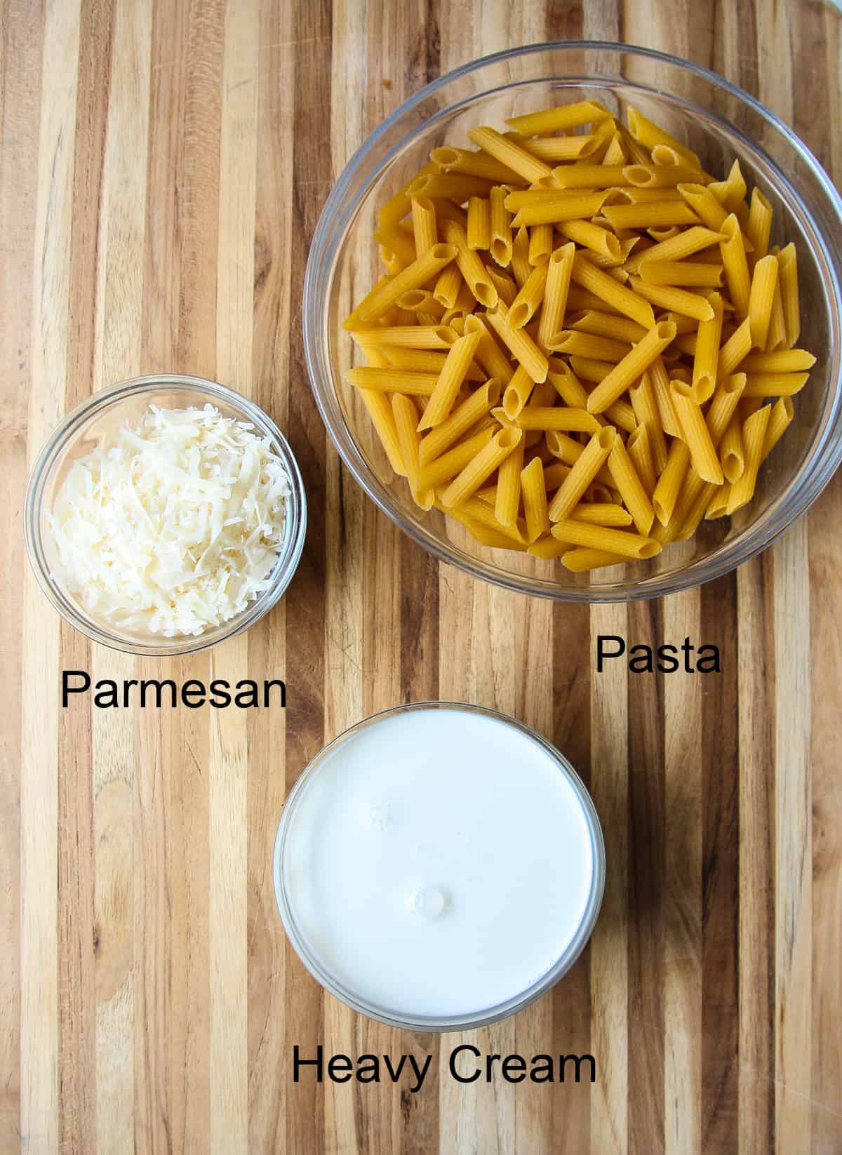 Uncooked penne pasta, grated parmesan cheese, and cream in glass bowls on a wooden board.